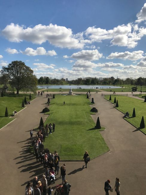 A wide view of Crystal Palace Park shows a well-maintained lawn with neatly trimmed grass and symmetrical flower beds adorned with small, conical-shaped evergreen trees. The paved walkways on either s